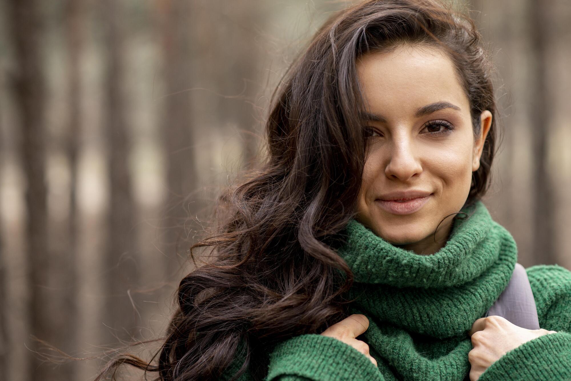 Mulher de cabelos longos, ondulados e escuros ao ar livre no inverno, usando cachecol de tricô verde, com fios soltos ao vento e fundo desfocado de árvores.