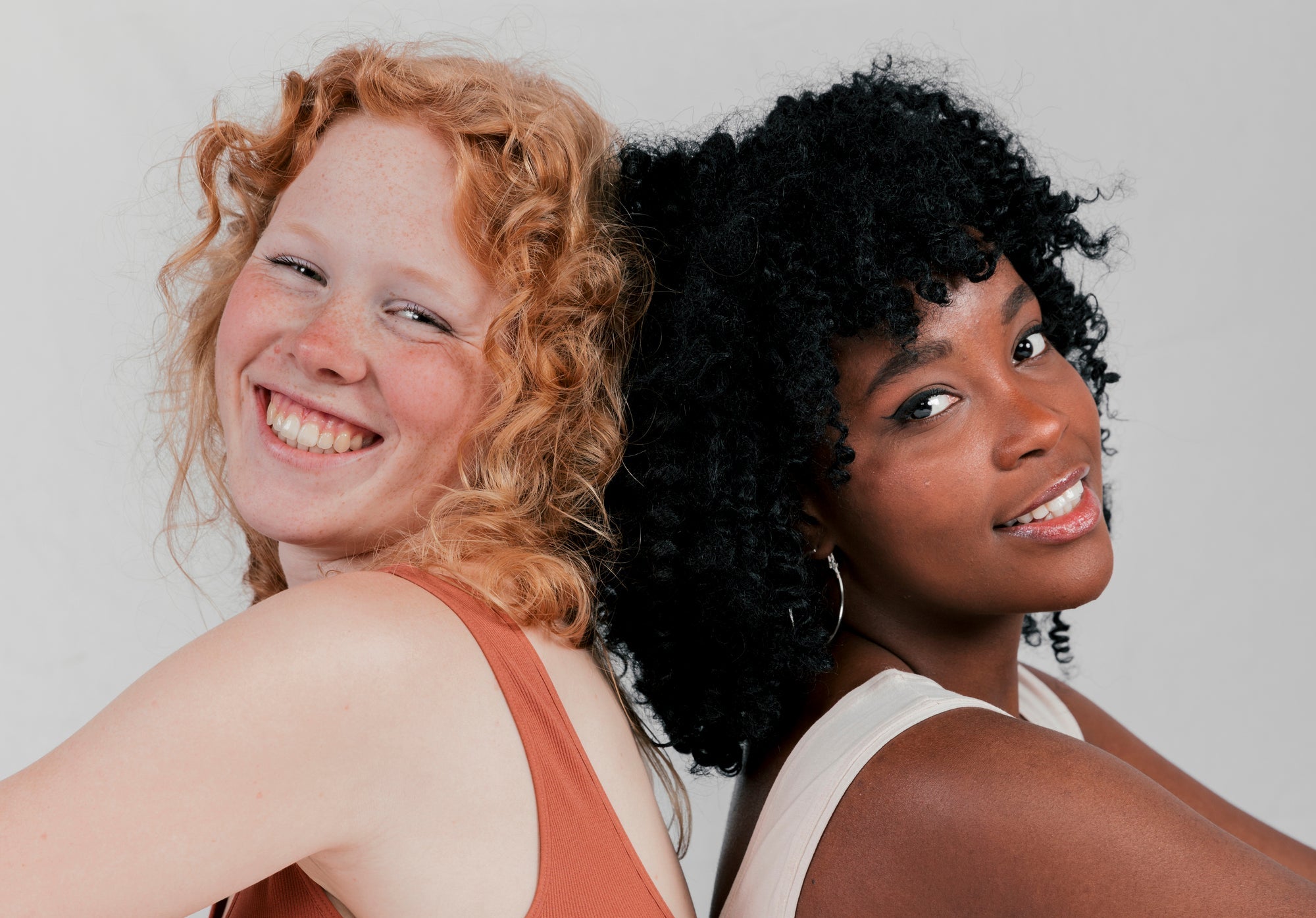 Duas mulheres jovens sorrindo lado a lado, uma de pele clara com cabelo ruivo cacheado e sardas e outra de pele negra com cabelo crespo, representando diferentes fototipos e tons de pele.