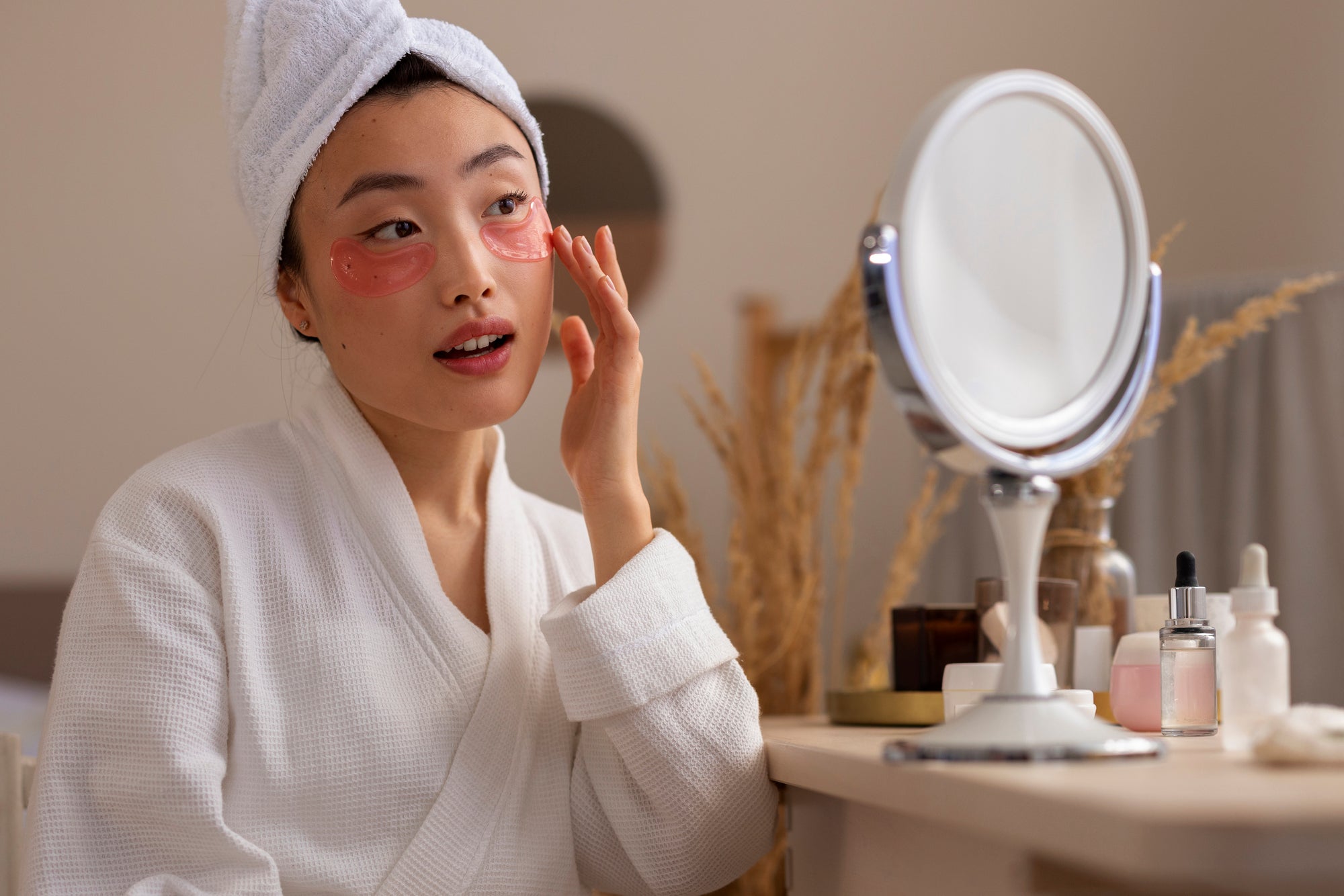 Mulher de roupão e toalha na cabeça, com máscaras para a área dos olhos, aplicando skincare em frente ao espelho de mesa com séruns, em uma rotina k-beauty.