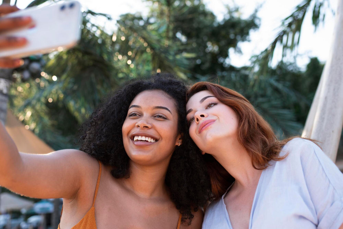 Duas mulheres de tons de pele diferentes sorriem em uma selfie ao ar livre, sob o sol e entre palmeiras.